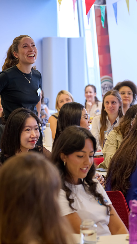 A woman standing up and smiling in a room of people who are sitting at tables.