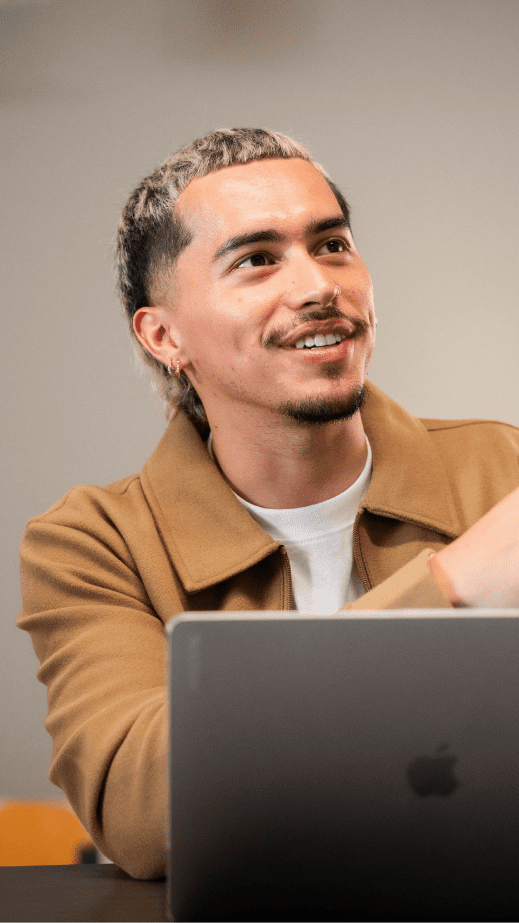 A man in a camel jacket sat smiling at a table with a laptop in front of him, looking to the right.